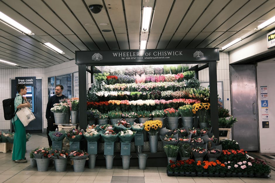 A vibrant floral display at Wheeler of Chiswick inside an underground station, featuring a variety of fresh flowers arranged in multiple tiers. The arrangement includes colorful roses, sunflowers, tul