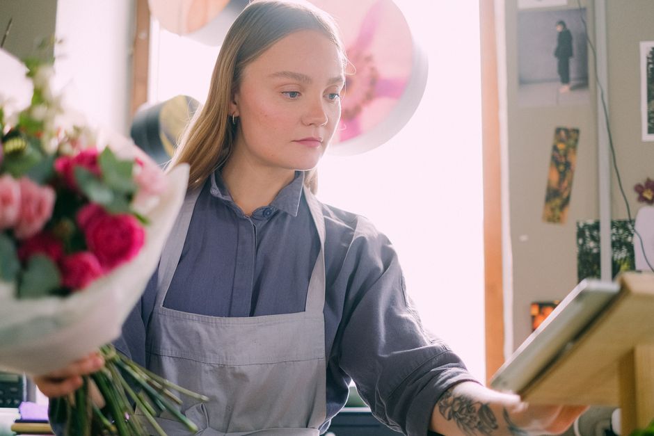 A young female florist with long, light brown hair wearing a gray apron over a blue shirt, carefully arranging a bouquet of fresh flowers with pink roses, white lilies, and greenery wrapped in white p