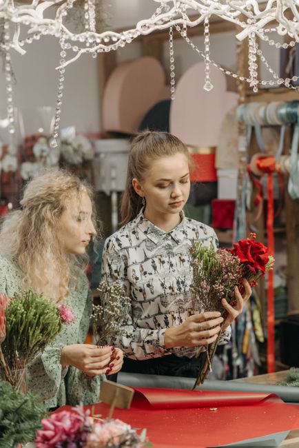 A young woman with long, light brown hair tied back, holding a bouquet of mixed flowers including red roses and purple statice, arranged with greenery and wrapped in clear cellophane, is examining the