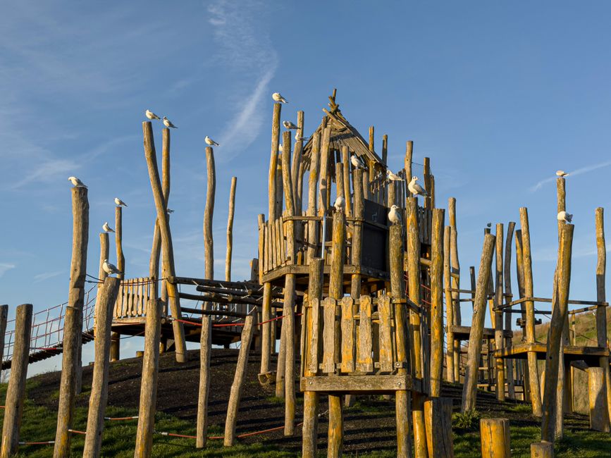 The image depicts a whimsical wooden play structure built from irregularly shaped logs and planks, resembling a treehouse or fort, situated outdoors on a grassy slope. The structure features multiple 
