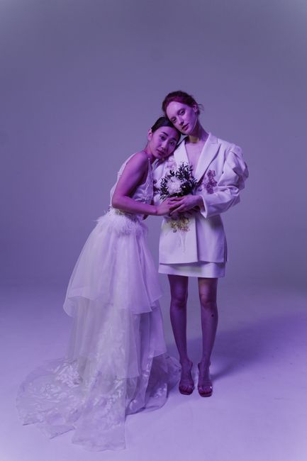 Two women pose together against a plain background, with one holding a floral gift. The woman on the left wears a delicate, flowing white dress with a sheer, layered tulle skirt and intricate lace det