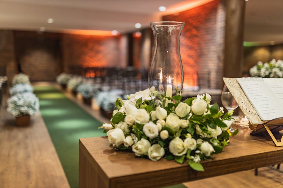 A close-up view of a floral arrangement featuring a bouquet of fresh white roses with lush green foliage, arranged around a clear glass hurricane lantern with a lit candle inside. The bouquet is place