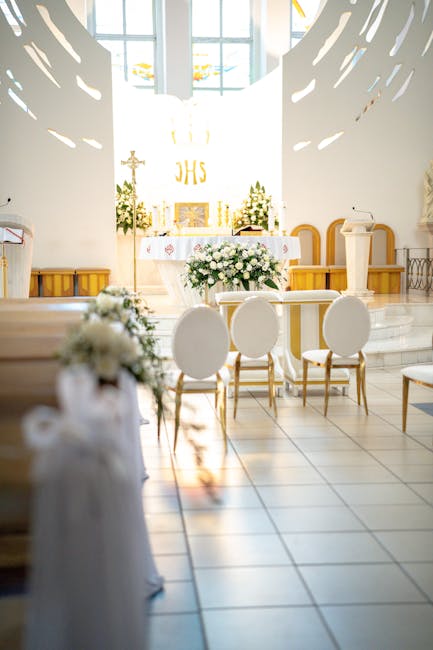 A floral arrangement featuring a bouquet of white roses, lilies, and greenery, displayed in a glass vase with a satin ribbon, placed on a table in the church interior. The arrangement is fresh, with v