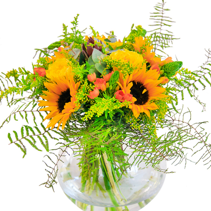 At first glance, this bouquet is pure radiance poured into floral form-a celebration of Hillingdon's bright and cheerful personality. Lustrous sunflowers with deep brown hearts and sunny petals stretch outward, basking in imagined sunlight reminiscent of afternoons spent in Fassnidge Park. Interspersed are elegant, buttery yellow roses, their petals unfurling in intricate spirals that catch the eye and conjure the scent of beloved English rose gardens. Ruby-red Hypericum berries gleam between the blooms, their polished skins providing playful contrast to the matte softness of surrounding petals. Wisps of fern fronds cascade outward, weaving a story of untamed summer meadows and echoing the movement of the wind through local vine-covered lanes. Delicate green fillers, goldenrod's tiny yellow blossoms, and slender green flower spikes add further texture, while subtle eucalyptus leaves introduce a cooling, silvery-green hue. All gathered in a pristine, rounded glass vase, the vibrant stems form a lush foundation, their pattern visible through the shimmering water. Every element, from petal to leaf, is illuminated in clean light, crafting a bouquet that is lavish, aromatic, and inviting. Ideal for birthdays, special occasions, or simply to share happiness, this arrangement captures the lively charm found throughout Hillingdon-a perfect gesture to brighten any moment or home.