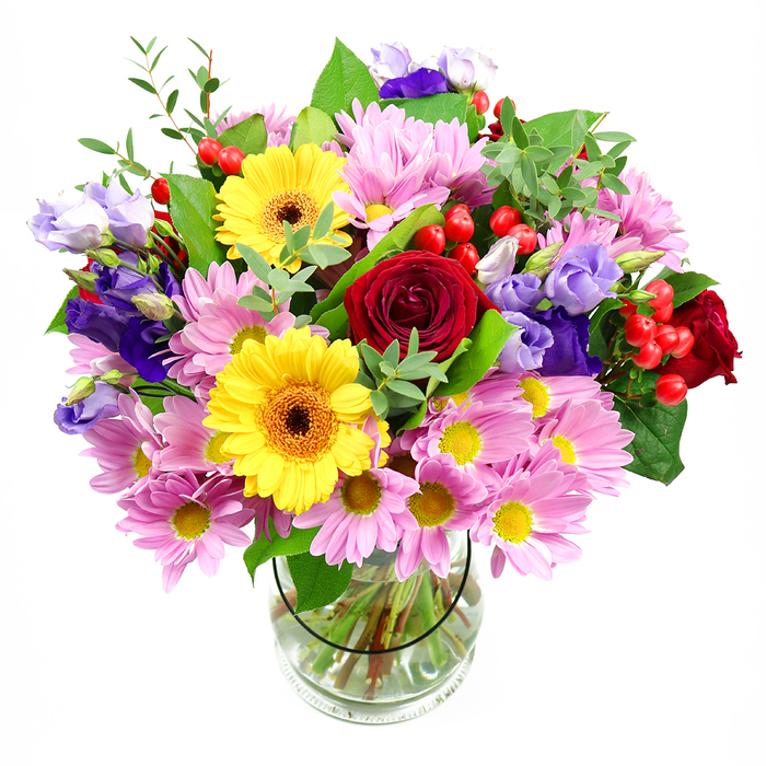 A generous bouquet of mixed blooms stands tall in a clear glass vase, its reflection softly visible against a white background that lets each flower's colour and form shine, much like the light-filled interiors of homes across Hillingdon. At the heart of the arrangement, sumptuous deep red roses bloom in velvety spirals, adding emotional depth and a timeless romantic note. Close by, two striking yellow gerbera daisies with rich brown-orange centres open like radiant suns, their broad petals creating a bold contrast that immediately draws the eye. Surrounding these focal blooms, clusters of soft pink and lilac daisies or chrysanthemums gather in abundance, each tiny flower formed of multiple slender petals circling a small yellow centre, building a textured cushion of pastel tones. Interlaced through these are stems of lisianthus in elegant shades of purple and lavender, their bell-shaped flowers and swelling buds introducing vertical lines and a sophisticated coolness, reminiscent of evening light settling over nearby parks and green spaces. Small, round hypericum berries in vivid red are dotted thoughtfully throughout the bouquet, like polished beads nestled between petals, lending a modern, tactile twist. A rich collar of greenery supports and frames the design, combining broad, dark leaves for structure with lighter, more delicate sprigs that soften the edges and give the impression of a hand-gathered garden posy. The transparent vase reveals neatly trimmed stems drinking from clear water, a quiet detail that speaks to professional care and freshness. The overall feel is joyful yet composed, a perfect floral gift for birthdays, anniversaries, or thank-you gestures delivered anywhere from Hillingdon Village to the quieter streets off Long Lane, where a splash of colour can instantly lift the atmosphere of a room.